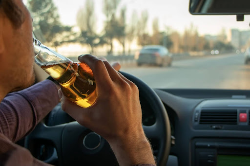 A man is actively drinking a beer while driving a car. The photo is from the backseat so you can not see his face. He is a danger to everyone.