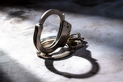 A pair of steel handcuffs is pictured. The background is grayish white. A shadow of the handcuffs stretches down to the lower right of the image.